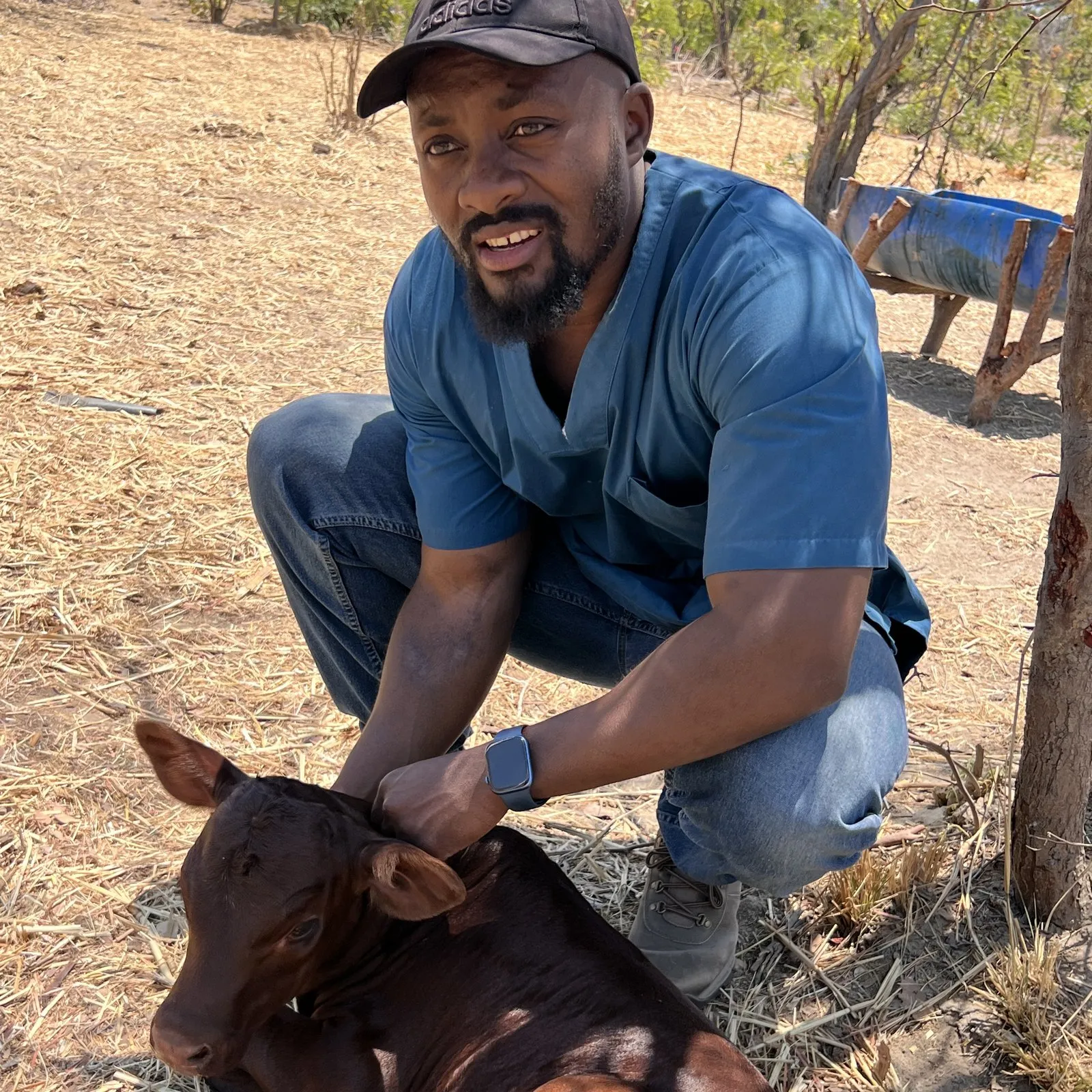 Muhuma M. crouching beside a calf at the farm, one hand resting on its head.